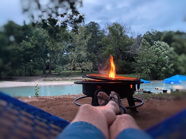 A person lying on a hammock with a grill in the foreground, a body of water, and trees in the background.