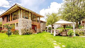 A modern house with a stone facade, large windows, and a well-maintained garden with a gazebo and outdoor seating. - Comfortable Villa in the Sacred Valley: Hot Tub, Sauna, Views of the Waterfall and Mountains (Calca)