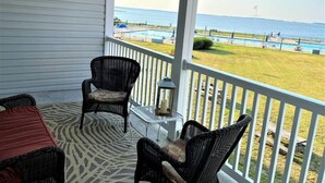 A covered balcony with wicker chairs and a table, overlooking a beach and a pool.