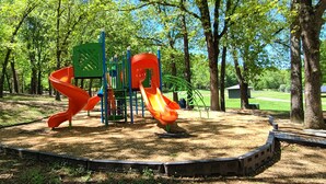 Une aire de jeux avec deux toboggans et une structure d’escalade, le tout entouré d’arbres.