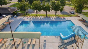 Une grande piscine rectangulaire avec un toboggan, entourée de chaises longues et de parasols, aménagée dans un espace paysager avec des arbres et un parking en arrière-plan.