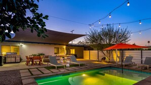 Un jardin avec une piscine, du mobilier d’extérieur, un espace barbecue et des guirlandes lumineuses.