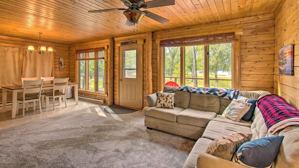 A cozy cabin interior with a wooden ceiling, a ceiling fan, a sofa, a dining table with chairs, and large windows offering a view of the outdoors. - Cozy Riverfront Getaway: Fishing On-Site! (Lacon)