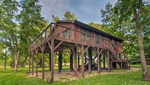A rustic wooden cabin with a balcony, situated in a wooded park area. - Cozy Riverfront Getaway: Fishing On-Site! (Lacon)