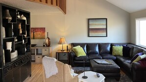 A modern living room with a black leather sofa, a wooden staircase, a ceiling fan, a black cabinet with shelves, a painting, and a window.