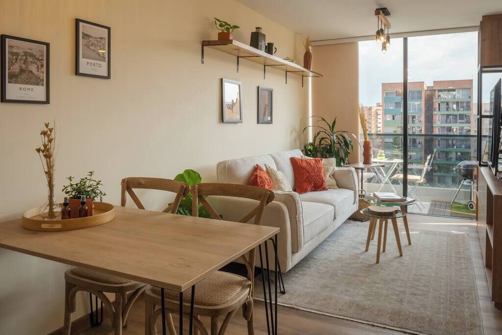 Una sala de estar moderna con un sofá, una mesa de comedor y sillas. Hay cuadros enmarcados en la pared, una planta en un estante y vistas de edificios a través de amplias ventanas.