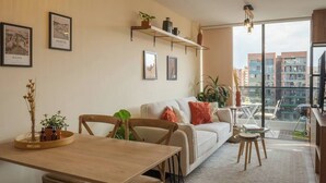 A modern living room with a sofa, a dining table, and chairs. There are framed pictures on the wall, a plant on a shelf, and a view of buildings through large windows. - Organic, Comfy & New loft in Laureles Quarter (Medellín)