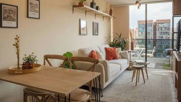 Una sala de estar moderna con un sofá, una mesa de comedor y sillas. Hay cuadros enmarcados en la pared, una planta en un estante y vistas de edificios a través de amplias ventanas.
