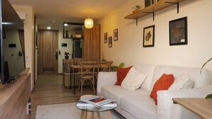 A modern living room with a white sofa, a coffee table, and a TV on a wooden stand. There is a dining area with a wooden table and chairs. - Organic, Comfy & New loft in Laureles Quarter (Medellín)