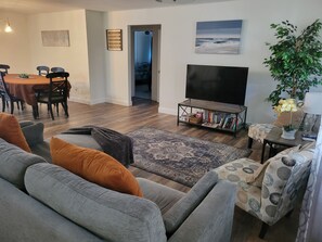A living room with a sofa, a coffee table, a TV stand with a flat-screen TV, a dining area with a table and chairs, and a hallway leading to another room.