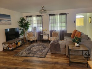Una sala de estar con televisor, dos sillones, un sofá, una mesa de centro, un tapete y una planta en maceta.