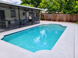 Una piscina rectangular con borde de azulejos, un porche cubierto con mobiliario exterior y una valla de madera al fondo.