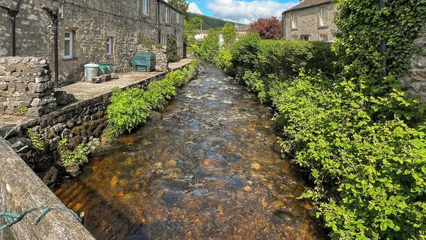 A narrow water channel between stone buildings with greenery on the sides.