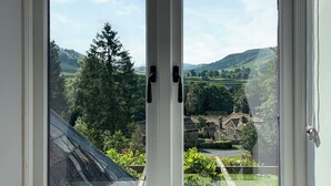 A view through a window with a stone wall and greenery, including trees and a distant village.