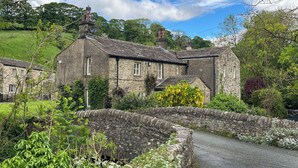 A stone house with a stone bridge in front, surrounded by greenery.