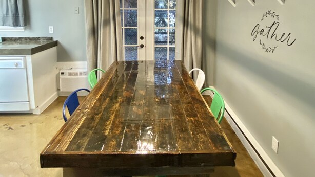 Un comedor con una mesa de madera, sillas, una lámpara colgante y una cocina al fondo.