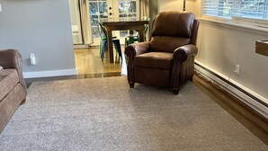 Una sala de estar con un sillón de cuero marrón, una mesa de comedor con sillas, una lámpara y una ventana con persianas.