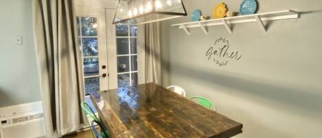 A dining area with a wooden table, colorful chairs, a pendant light, and a wall with decorative plates and a 'Gather' sign.