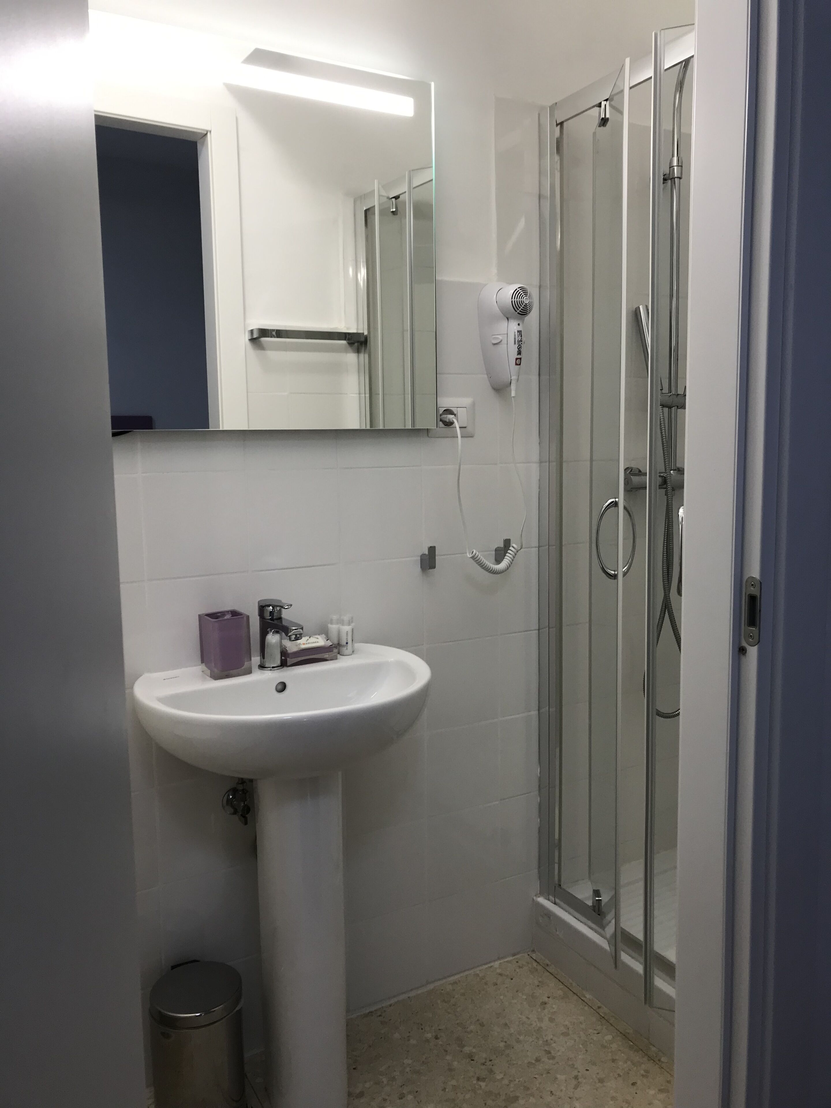 Un bagno con lavandino bianco, uno specchio, box doccia in vetro e un cestino.