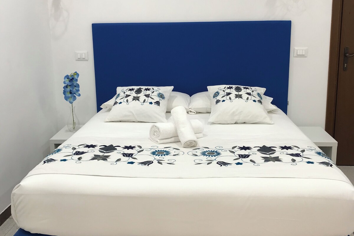 Un lit bien fait, avec une tête de lit bleue, une literie blanche et des coussins décoratifs. Il y a deux tables de chevet, une de chaque côté du lit, et une petite plante en pot est posée sur la table de gauche.