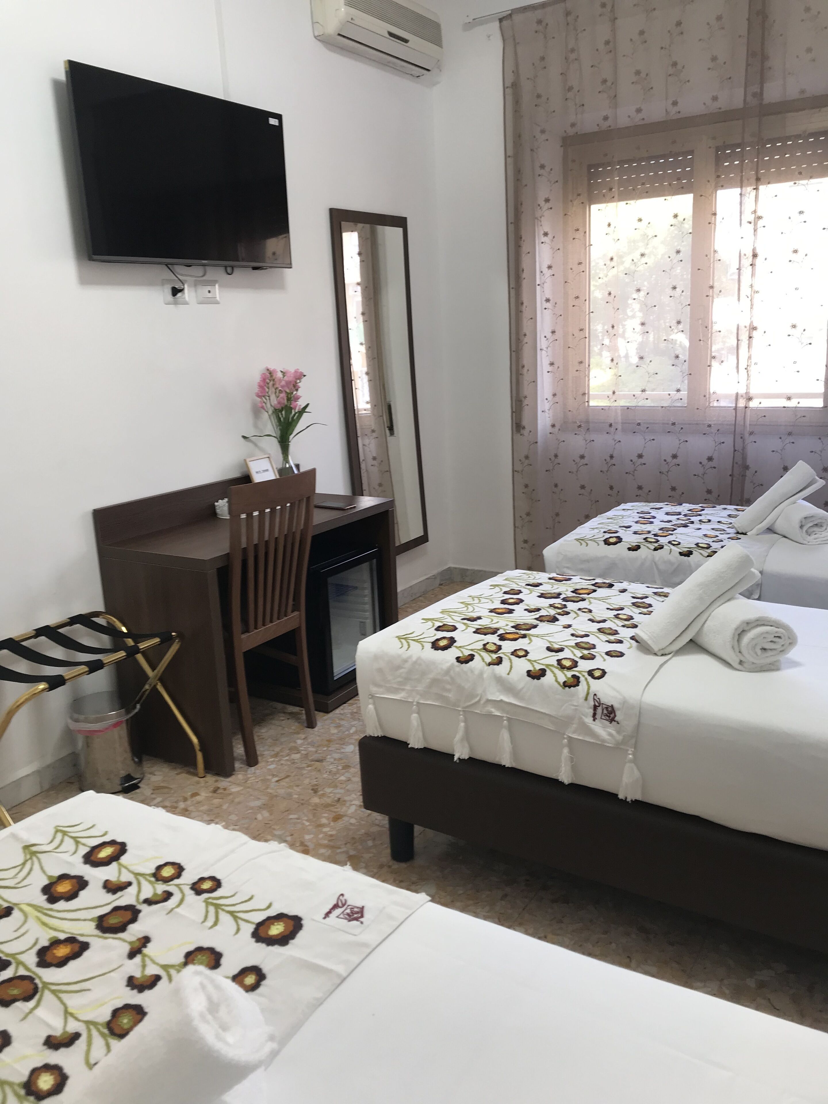 A hotel room with a bed, a desk, a chair, a TV, and a window with floral curtains.