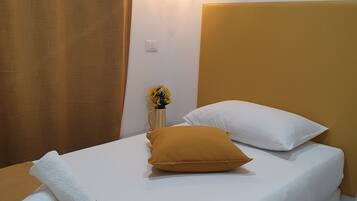 Una cama bien hecha con ropa de cama blanca y mostaza, un jarrón pequeño con flores amarillas y una lámpara colgante.