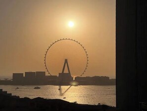 Uma roda gigante silhuetada contra o pôr do sol, com um corpo d'água ao primeiro plano.
