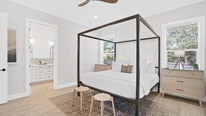 Ein modernes Schlafzimmer mit Himmelbett, Deckenventilator aus Holz, Nachttisch, Kommode und Blick ins Freie durch ein Fenster.