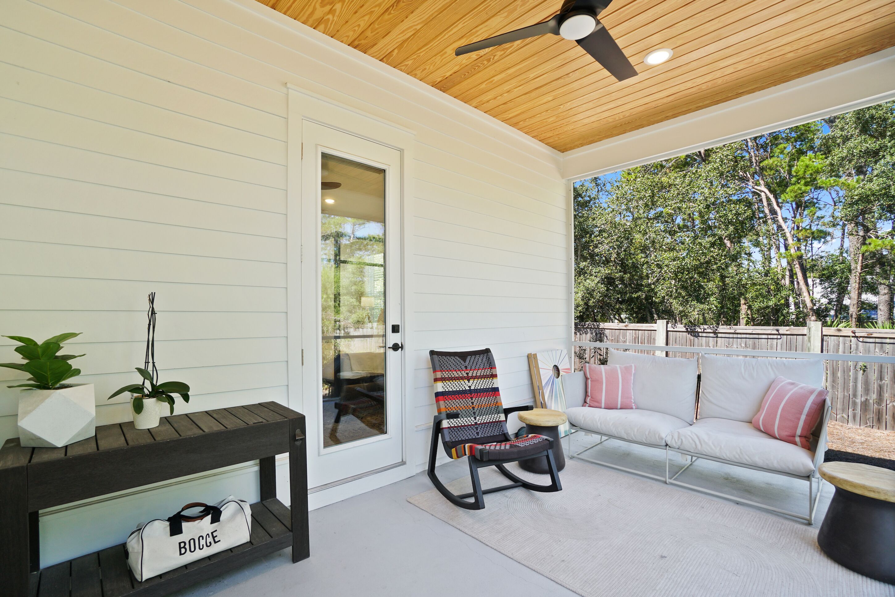 Un patio couvert avec un plafond en bois, un mur blanc, un banc sur lequel est posé un sac étiqueté « BOCCE », une chaise à bascule et un canapé blanc agrémenté de coussins roses.