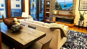 Een moderne woonkamer met een open haard, een eettafel en een bank. Er is uitzicht op een balkon en op de stad door de ramen.
