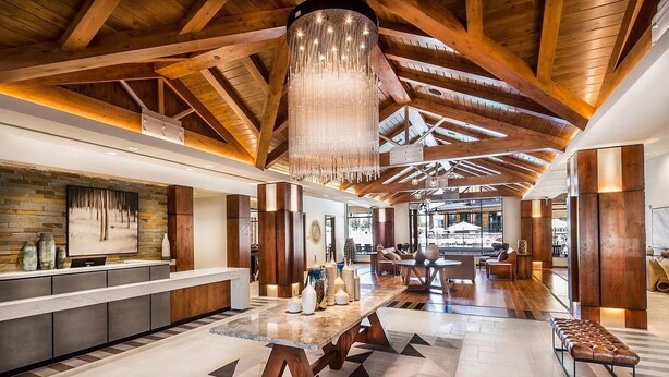 Een ruime lobby met een hoge plafond, houten balken, een centrale kroonluchter, een receptiebalie en een zithoek met uitzicht op de omgeving.