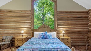 Un dormitorio de cabaña de troncos con un ventanal grande, una cama de madera, dos mesitas de noche con lámparas y una silla.