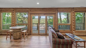 Una habitación amplia con detalles de madera, ventanas grandes, una mesa redonda de madera con taburetes, un sofá a rayas y una mesita con una revista.