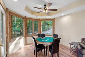 A room with a green felt poker table, wooden chairs, a ceiling fan, and large windows offering a view of trees.