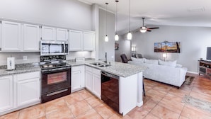 A modern kitchen with white cabinets, a built-in oven, and a dishwasher. There is a dining area with a white sofa and a ceiling fan. - Gilbert Family Home: Games, Private Pool & Patio! (Gilbert)