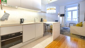 A modern kitchen with a built-in oven, a coffee machine, and a kettle. There is a small island with a stool, and a dining area with a sofa and a floor lamp. - Central Downtown Apartment by LovelyStay (Lisboa)