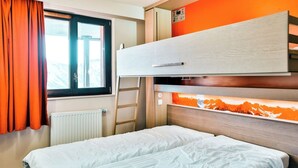 Ein Etagenbett mit massivem Kopfteil und einer unteren Liegefläche, ein Fenster mit orangefarbenen Vorhängen und ein Heizkörper.