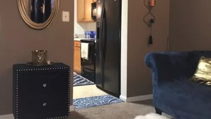 Una sala de estar con un sofá azul, un mueble negro, un espejo y una cocina al fondo.