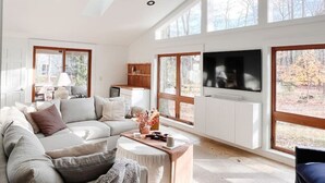 Ett rymligt vardagsrum med en stor soffa, ett soffbord och en tv. Det finns stora fönster som släpper in naturligt ljus och erbjuder utsikt över omgivningarna.
