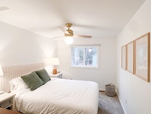 Une chambre à coucher comprenant un lit, une table de chevet, une lampe, un ventilateur de plafond, une fenêtre et des tableaux encadrés accrochés au mur.