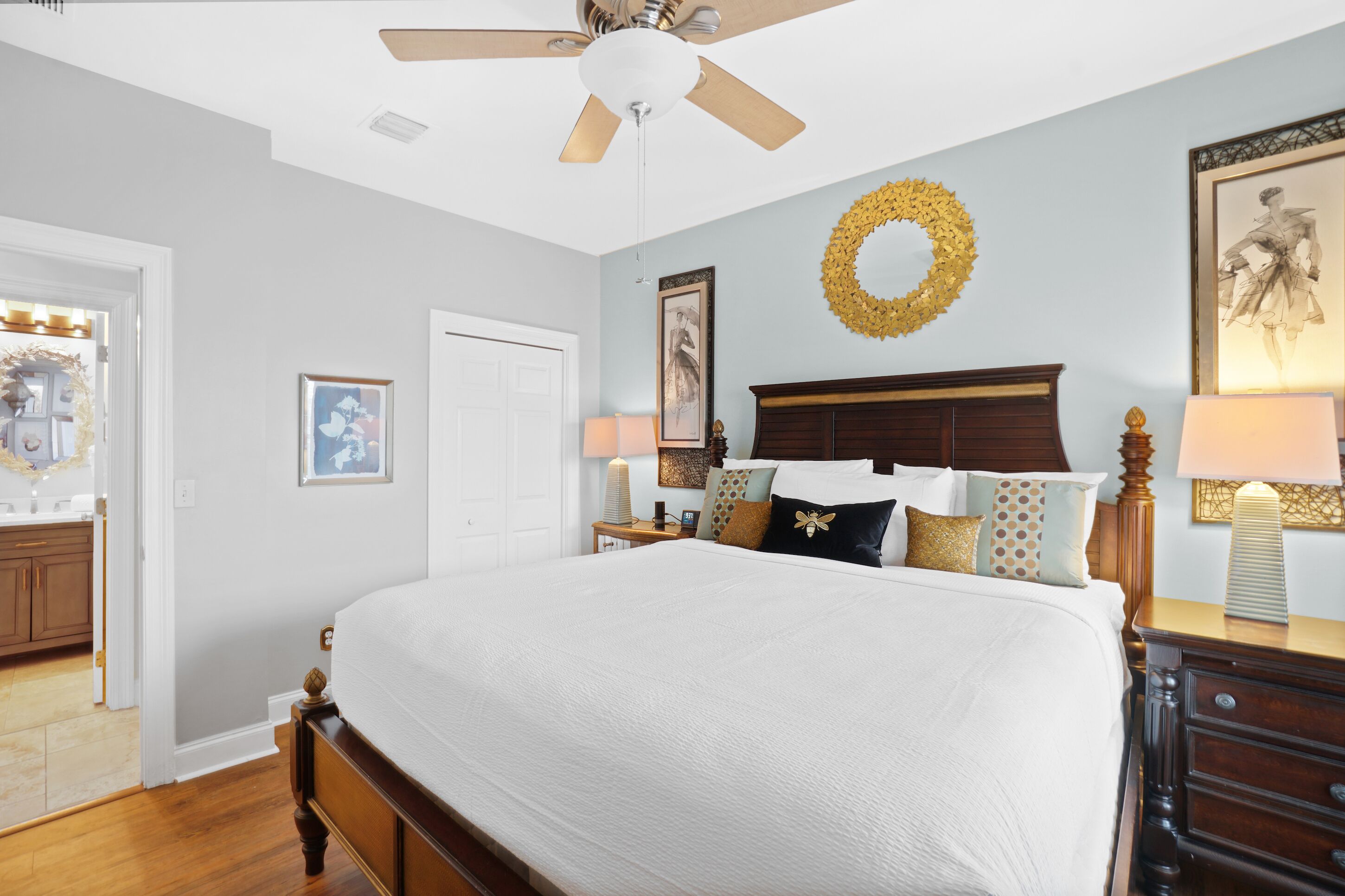 A bedroom with a large bed, a ceiling fan, a wooden headboard, a nightstand with a lamp, and a framed picture on the wall.