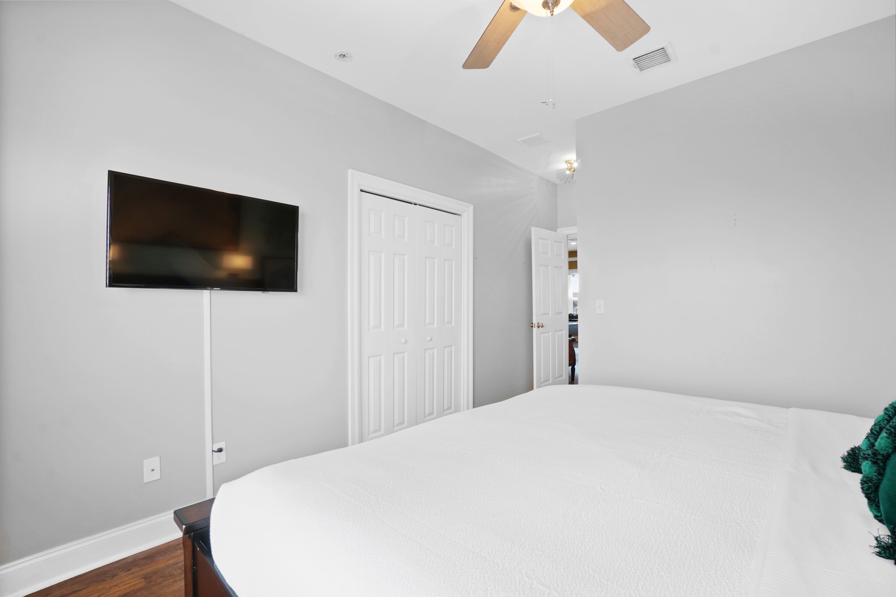 A bedroom with a bed, a TV mounted on the wall, a ceiling fan, and a door leading to another room.