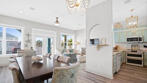 A spacious dining area with a large wooden table and patterned chairs, a kitchen with teal cabinets and a microwave, and a ceiling fan. - Beach House - Dreams Come True by PHG (Panama City Beach)