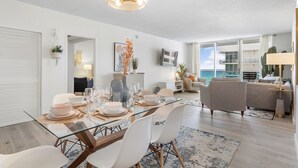 A modern living room with a glass dining table, white chairs, and a large window offering a view of the ocean. - Stunning ocean front unit 2/2 with privet beach excess (Hollywood)
