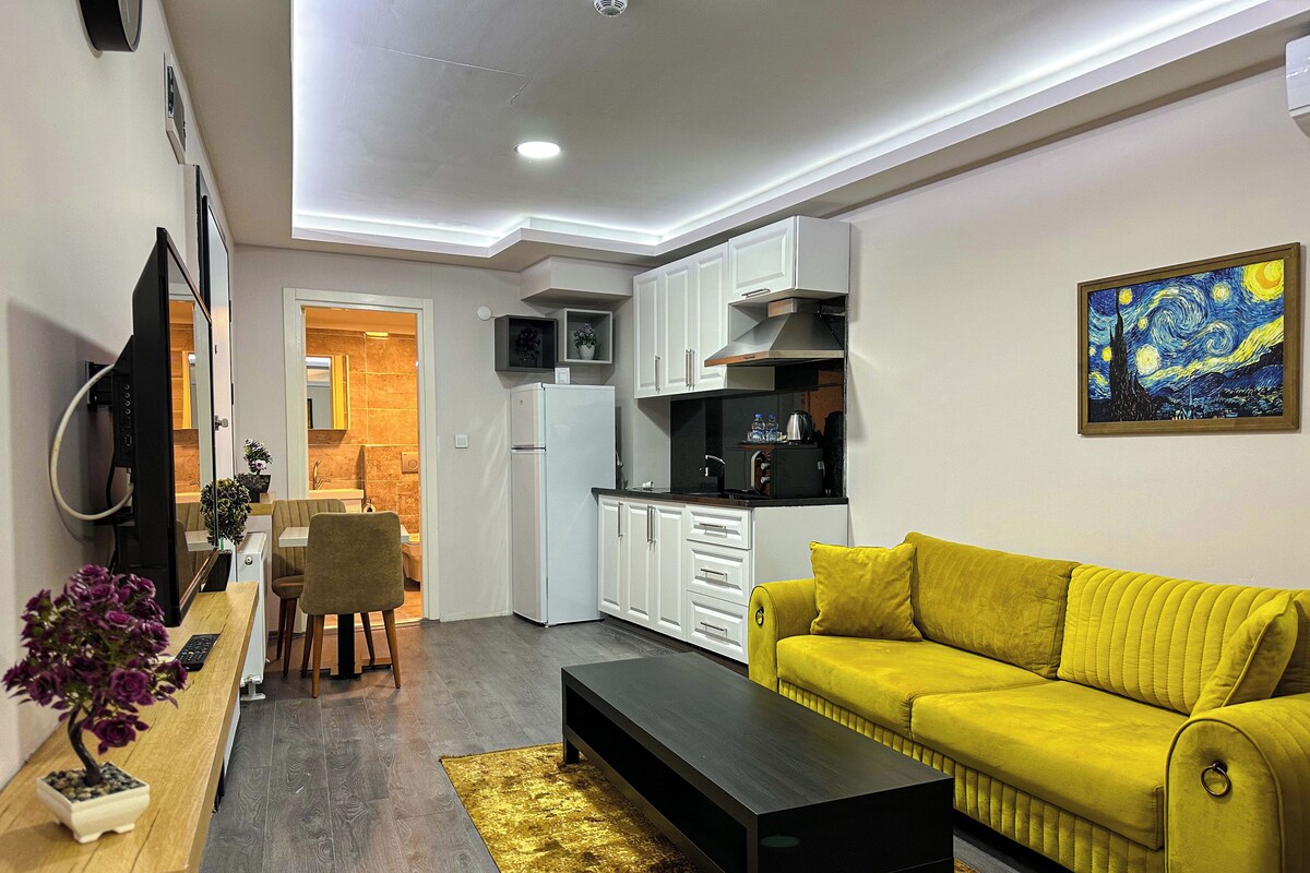 A modern living room with a yellow sofa, a black coffee table, and a kitchen area with white cabinets and a black countertop.