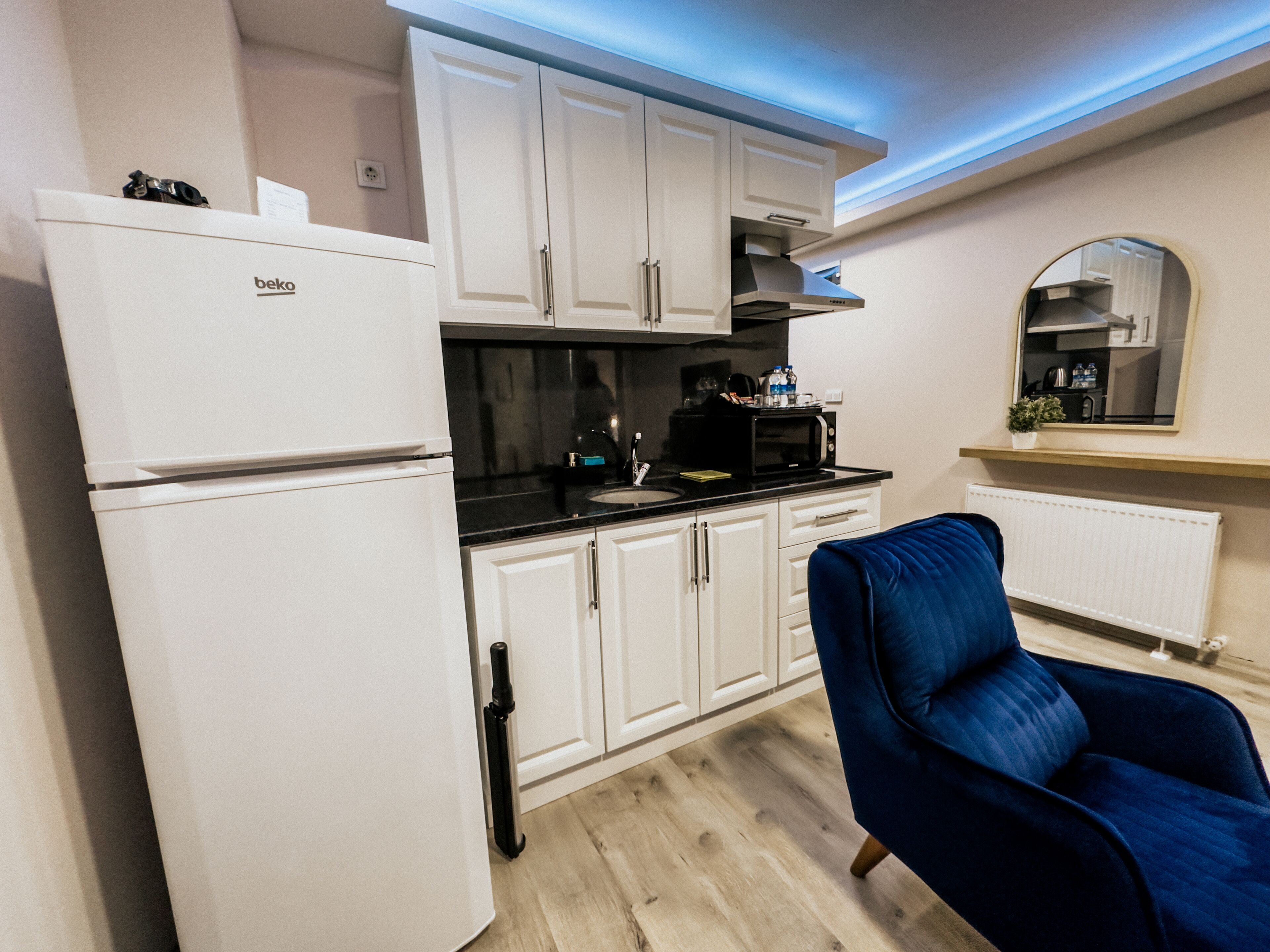 A compact kitchen with white cabinets, a black countertop, a white refrigerator, a blue chair, and a mirror above the counter.
