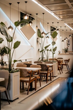 Un espace de restauration moderne, doté de plusieurs tables et chaises, d’un éclairage suspendu et de grands pots de plantes.
