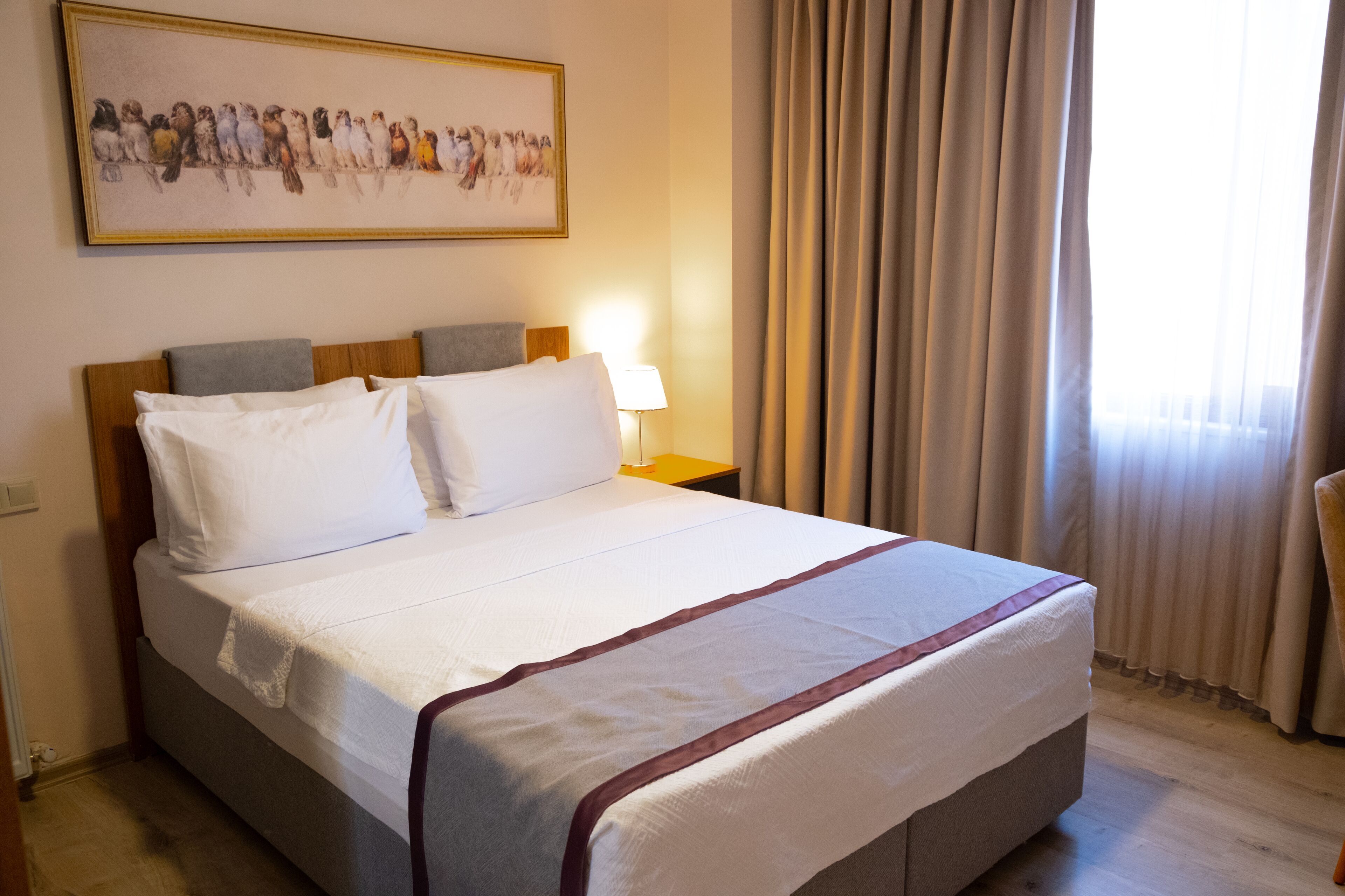 A neatly made bed with white linens and a red accent, a bedside lamp, and a framed bird painting on the wall.