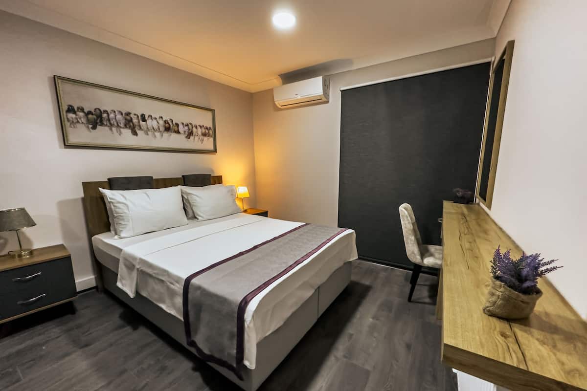 Une chambre à coucher moderne, comprenant un lit, des tables de chevet, un bureau, une chaise, une œuvre d’art encadrée et un appareil de climatisation.