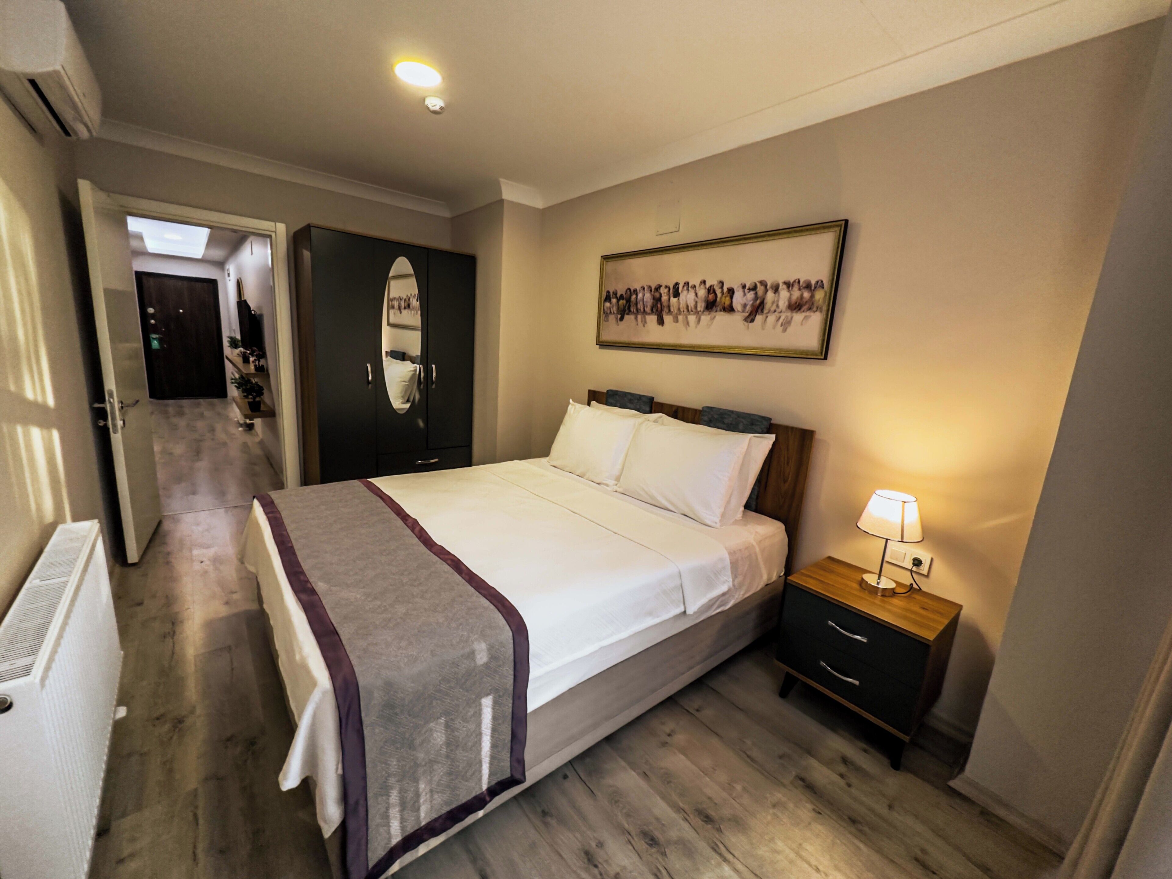 A hotel room with a bed, bedside table, lamp, wardrobe, and a hallway leading to another room.